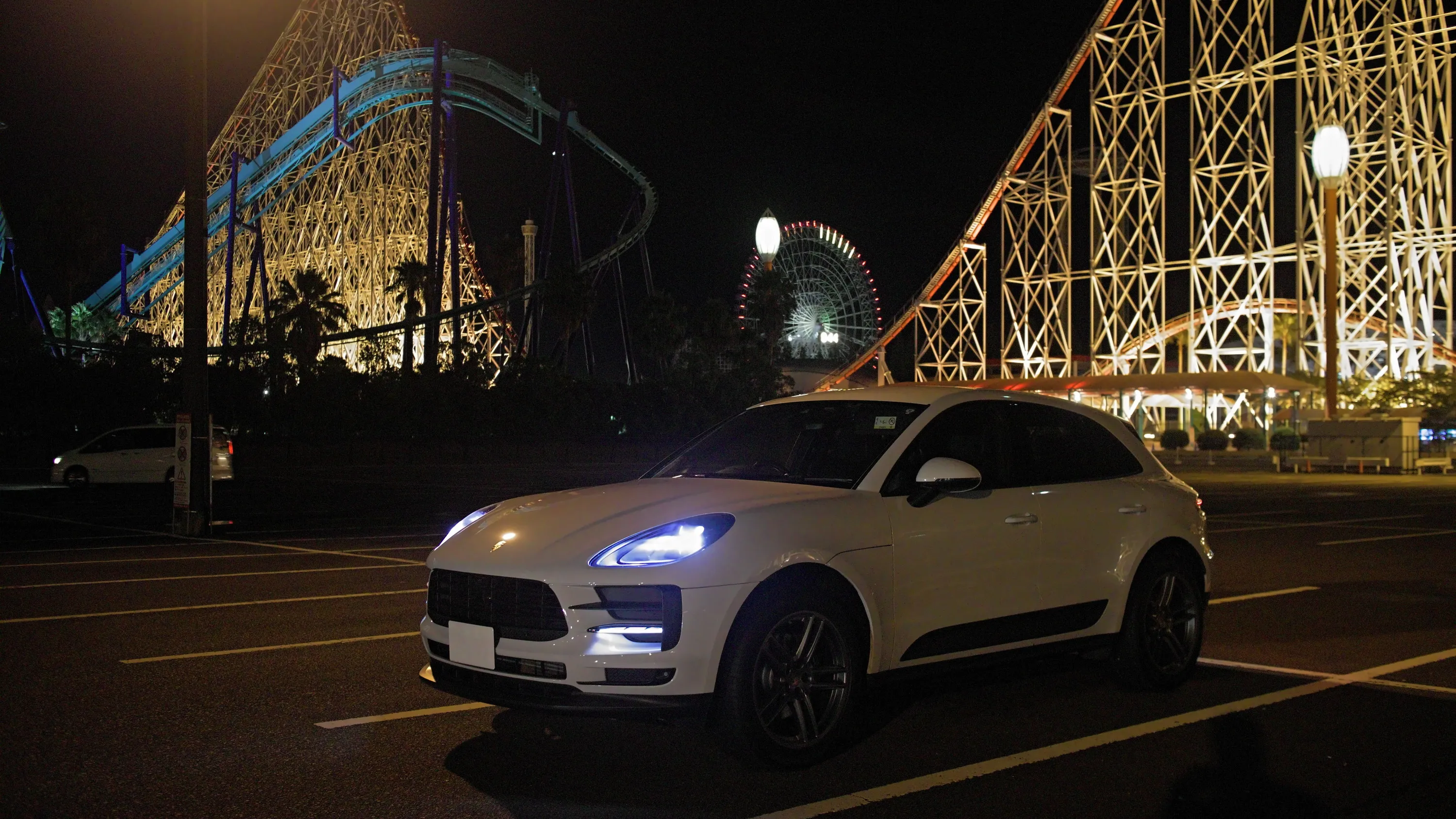 夜の遊園地の駐車場に停まる白いポルシェ・マカン。背後にライトアップされたジェットコースターと観覧車